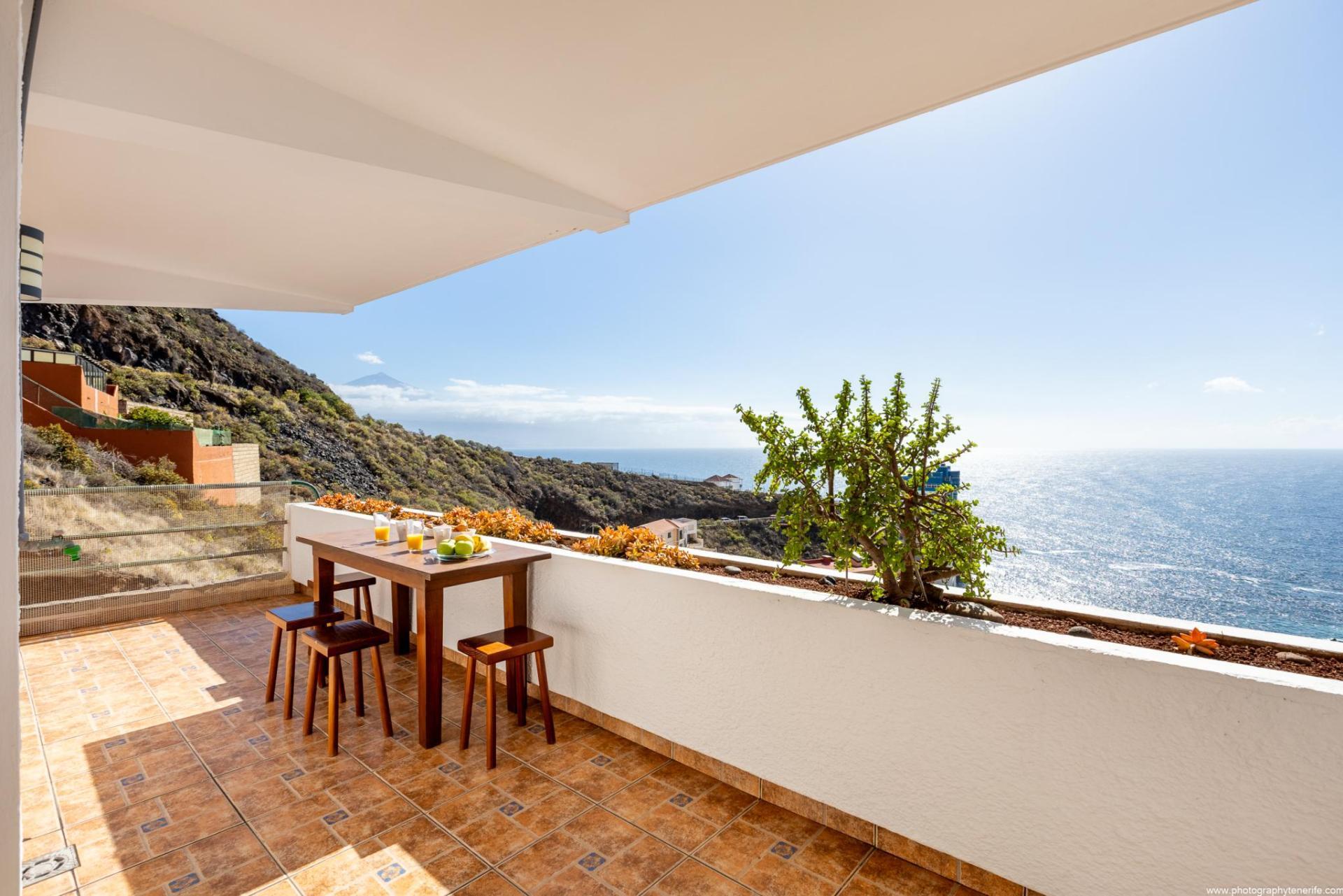 Alquiler vacacional canarias, balcón con mesas y sillas frente al mar azul, cielo claro despejado, montañas y vegetación alrededor.